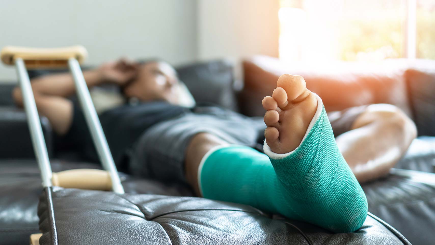 Man laying beside crutches on a couch with leg in a green cast recovering at home from a work injury